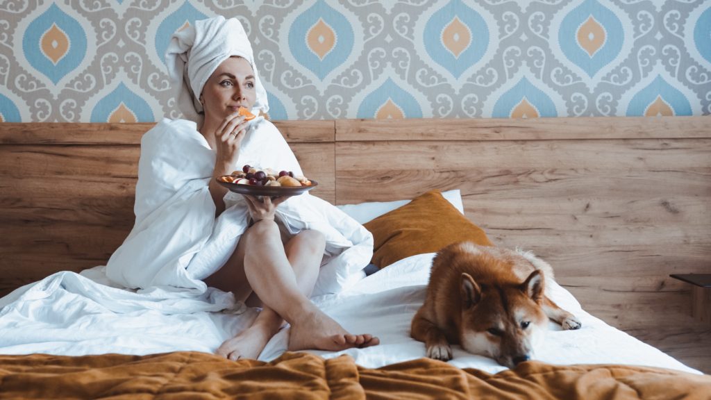 A girl with a towel on her head and a dog on the bed eats fruit for breakfast. Treat yourself